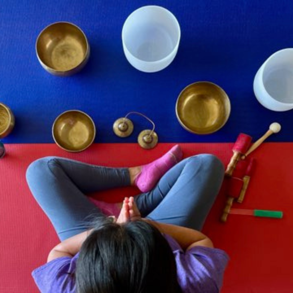 overhead image of Dorothy seated on yoga mats, hands at heart in greeting, with singing bowls in front on the floor